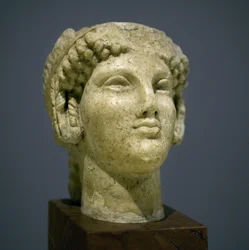 Testa di ragazza (Kore), scultura del periodo ellenistico romano (III-II secolo d.C.), volto molto espressivo, zigomi prominenti a est, sorriso, capelli ricci, stile arcaico di Ionia nel VI secolo a.C. che si perpetuò a lungo