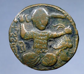 Dritto della moneta (bronzo) raffigurante un turco elmetto seduto a gambe incrociate che tiene una spada dietro la testa nella mano destra e una testa mozzata elmetto nella mano sinistra. coniata da Husam al-Din Yuluq Arslan (1184-1201)