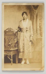 Cartolina fotografica di una donna accanto a una sedia di legno, 1904-1918