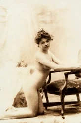 Ritratto nudo di una giovane donna inginocchiata davanti a una piccola poltrona.
