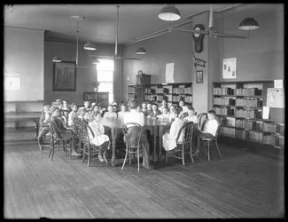 Bambini ascoltano una donna nella sala di lettura di una filiale non identificata della Biblioteca Pubblica del Queens Borough, c.1910