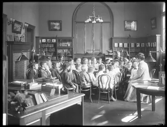 Bambini ascoltano una donna nella sala di lettura di una filiale non identificata della Biblioteca Pubblica del Queens Borough, c.1910