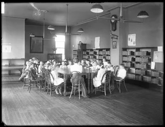 Bambini ascoltano una donna nella sala di lettura di una filiale non identificata della Queens Borough Public Library, ca. 1910