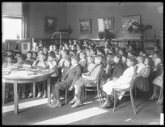 Bambini seduti, che ascoltano nella sala di lettura di una filiale non identificata della Biblioteca Pubblica del Queens Borough, c.1910