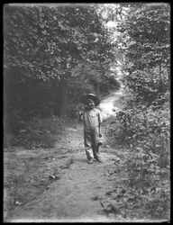 William Gray Hassler con cappello di paglia e salopette, che porta una canna da pesca e un cestino, camminando lungo un sentiero nel bosco