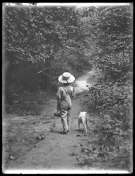 William Gray Hassler con cappello di paglia e salopette, che porta una canna da pesca e un cestino, camminando lungo un sentiero nel bosco con un piccolo cane (Bessie), 20 agosto 1911
