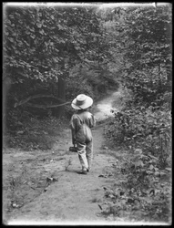 William Gray Hassler con cappello di paglia e salopette, che porta una canna da pesca e un cestino, camminando lungo un sentiero nel bosco da solo, 20 agosto 1911